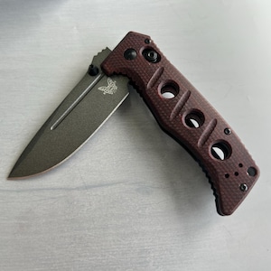 Può includere: Un coltellino tascabile rosso con una lama nera e un manico testurizzato. Il manico ha un logo a forma di farfalla e diversi fori.