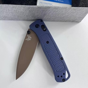 May include: A blue folding knife with a brown blade. The handle features a textured design and black fixtures. The blade is partially open, displaying a butterfly logo. The knife is set against a neutral background.