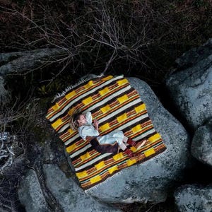 May include: Overhead shot of a person lying on a colorful blanket atop large gray rocks. The blanket features a striped pattern in yellow, brown, and white. The person is wearing light-colored clothing and brown boots.