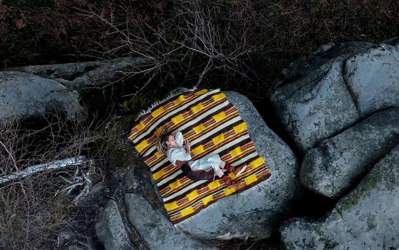 May include: Overhead shot of a person lying on a colorful blanket atop large gray rocks. The blanket features a striped pattern in yellow, brown, and white. The person is wearing light-colored clothing and brown boots.
