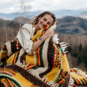 May include: A colorful, woven blanket with a mix of yellow, brown, red, and green stripes and accents. The blanket has a fringed edge and is adorned with decorative, textured elements. The blanket is wrapped around a person.