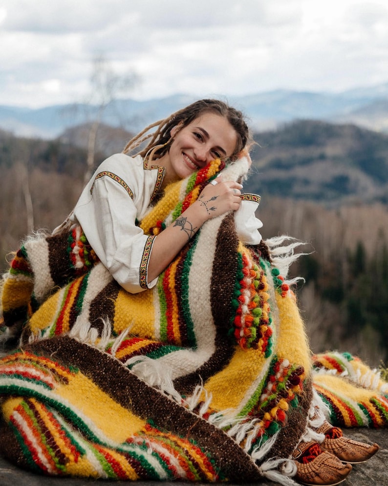 May include: A colorful, woven blanket with a mix of yellow, brown, red, and green stripes and accents. The blanket has a fringed edge and is adorned with decorative, textured elements. The blanket is wrapped around a person.