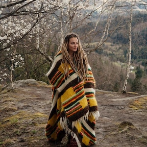May include: A colorful, striped poncho in shades of yellow, brown, and red. The poncho has a fringed edge and is worn outdoors. The person wearing the poncho has long dreadlocks and is standing on a rocky surface.