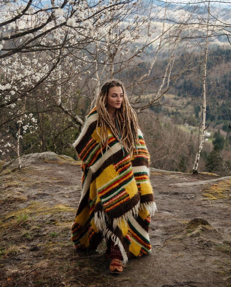 May include: A colorful, striped poncho in shades of yellow, brown, and red. The poncho has a fringed edge and is worn outdoors. The person wearing the poncho has long dreadlocks and is standing on a rocky surface.