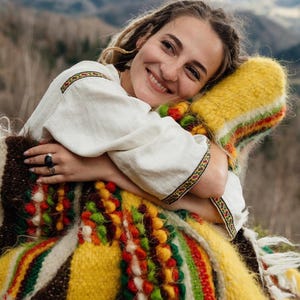 May include: A woman wearing a white embroidered shirt with colorful trim, embracing a vibrant, hand-woven blanket. The blanket features a mix of yellow, red, green, and brown hues, creating a textured, folk-art aesthetic. The background shows a blurred mountain landscape.