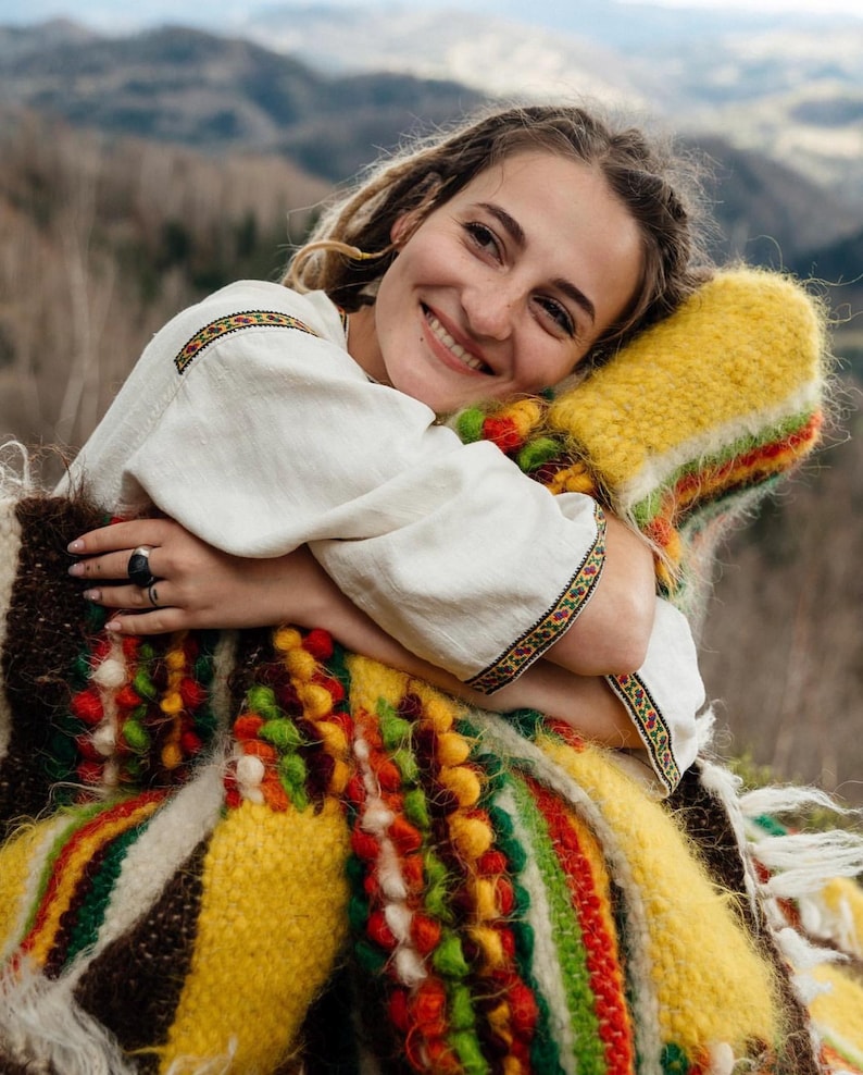 May include: A woman wearing a white embroidered shirt with colorful trim, embracing a vibrant, hand-woven blanket. The blanket features a mix of yellow, red, green, and brown hues, creating a textured, folk-art aesthetic. The background shows a blurred mountain landscape.