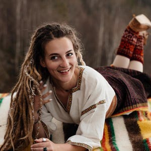 May include: A woman with long dreadlocks smiles while lying on a colorful blanket. She wears a white blouse with embroidered trim and a dark skirt. Her legs are covered with patterned leg warmers. The background is a blurred forest.