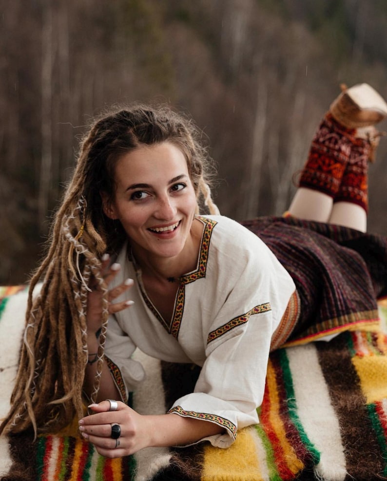 May include: A woman with long dreadlocks smiles while lying on a colorful blanket. She wears a white blouse with embroidered trim and a dark skirt. Her legs are covered with patterned leg warmers. The background is a blurred forest.