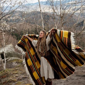 May include: A colorful woven blanket with a striped and geometric pattern. The blanket features shades of yellow, brown, red, and green. The blanket has a fringed edge and is being held up in the air.