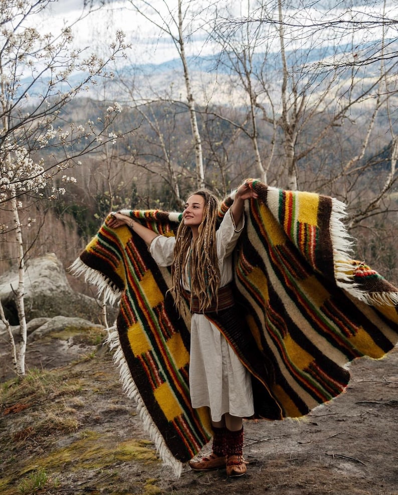 May include: A colorful woven blanket with a striped and geometric pattern. The blanket features shades of yellow, brown, red, and green. The blanket has a fringed edge and is being held up in the air.