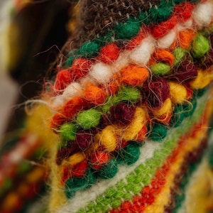 May include: Close-up of a vibrant, textured textile featuring a mix of colors. The design incorporates a variety of textures and hues, including red, orange, green, yellow, and white. The textile has a fuzzy, handmade appearance, suggesting a craft item.