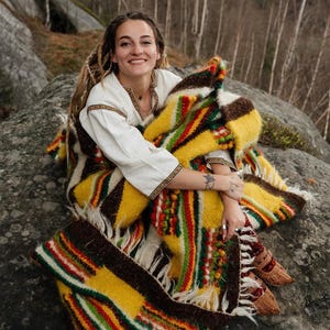 May include: A woman wearing a white embroidered shirt and a colorful woven blanket. The blanket features yellow, brown, red, and green stripes and fringe. The woman is sitting on a large rock outdoors.