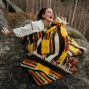 May include: A colorful, patterned blanket with yellow, brown, green, and red stripes. The blanket is draped over a person wearing a white shirt with embroidered details. The scene is set outdoors on a large rock.