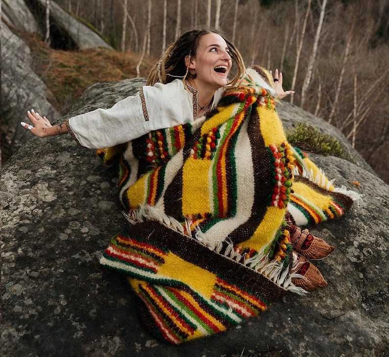 May include: A colorful, patterned blanket with yellow, brown, green, and red stripes. The blanket is draped over a person wearing a white shirt with embroidered details. The scene is set outdoors on a large rock.