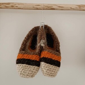 May include: A pair of brown, orange, and beige knitted slippers hanging on a wooden hanger. The slippers have a striped pattern and are made of wool.