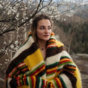 May include: A colorful, striped blanket in shades of yellow, brown, red, green, and white. The blanket is wrapped around a person, with a backdrop of a tree with white flowers and a blurred outdoor scene.