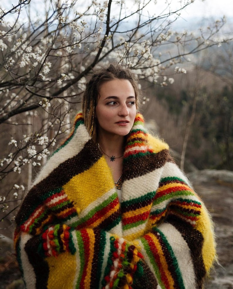 May include: A colorful, striped blanket in shades of yellow, brown, red, green, and white. The blanket is wrapped around a person, with a backdrop of a tree with white flowers and a blurred outdoor scene.