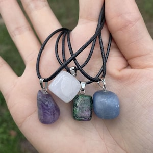 Könnte beinhalten: Vier verschiedene farbige Edelstein-Anhänger an schwarzen Schnüren. Die Steine sind Amethyst, Weiß, Grün und Blau.