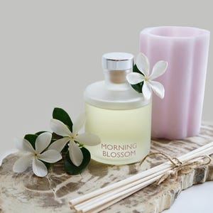 May include: Aromatherapy diffuser with the text "MORNING BLOSSOM", a light pink candle, and white flowers. The diffuser has a silver cap and is filled with a yellow liquid. Reed sticks are tied with twine. The items are on a decorative wooden slab.