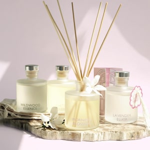 May include: Four glass diffuser bottles with silver caps and wooden reeds, labeled "Wildwood Essence," "White Tea," "Morning Blossom," and "Lavender Elixir." The bottles sit on a wooden slab with a pink background. A small gift box is also present.