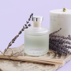May include: A frosted glass bottle with a silver cap and the words "LAVENDER ELIXIR" sits on a wooden slab, accompanied by a marble container and dried lavender sprigs. Reed diffuser sticks are tied with twine. The background is a soft purple.