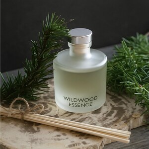 May include: A frosted glass diffuser bottle with a silver cap, labeled "WILDWOOD ESSENCE", sits on a stone slab. Bundled wooden reeds and sprigs of greenery frame the bottle, creating a natural, spa-like aesthetic. The background is a dark gray.