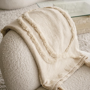 May include: Cream-colored woven throw blanket with textured fringe detail and tassels. The blanket is draped over a cream-colored chair, with the Elle Home logo visible in the upper right corner. The blanket's design features a textured, raised pattern.