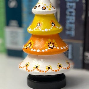 May include: A whimsical, hand-painted wooden tree figurine. The tree is composed of four tiers in white, yellow, and orange, adorned with small floral patterns. It sits on a black base, creating a decorative accent piece.