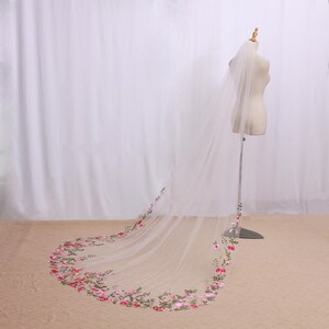 May include: A long, sheer white bridal veil with a floral embroidered border in shades of pink and green. The veil is displayed on a mannequin, showcasing its length and delicate design. The floral embroidery adds a touch of elegance.
