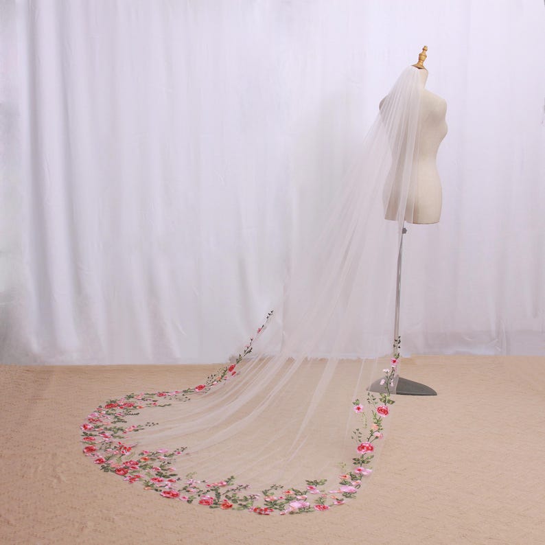 May include: A long, sheer white bridal veil with a floral embroidered border in shades of pink and green. The veil is displayed on a mannequin, showcasing its length and delicate design. The floral embroidery adds a touch of elegance.
