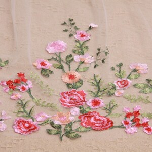 May include: Embroidered floral fabric featuring a delicate arrangement of pink and red flowers with green leaves. The flowers are of varying sizes and shapes, creating a textured and detailed design. The fabric has a soft, neutral background.