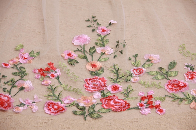 May include: Embroidered floral fabric featuring a delicate arrangement of pink and red flowers with green leaves. The flowers are of varying sizes and shapes, creating a textured and detailed design. The fabric has a soft, neutral background.
