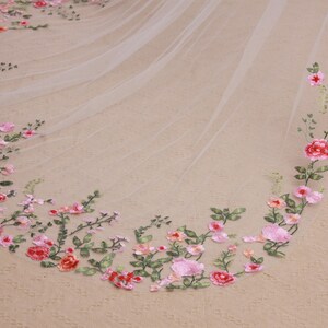 May include: A delicate, ivory-colored wedding veil with an embroidered floral design. The embroidery features pink and red roses, with green leaves and stems. The veil is made of a sheer, lightweight fabric.