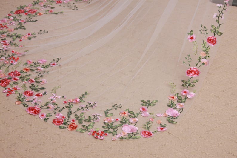 May include: A delicate, ivory-colored wedding veil with an embroidered floral design. The embroidery features pink and red roses, with green leaves and stems. The veil is made of a sheer, lightweight fabric.