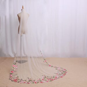 May include: A long, sheer white bridal veil with a floral embroidered border in shades of pink, red, and green. The veil is displayed on a mannequin, showcasing its length and delicate design. The veil's floral embroidery adds a touch of elegance.