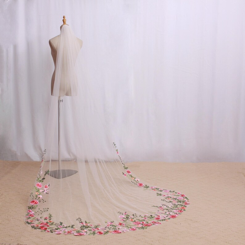 May include: A long, sheer white bridal veil with a floral embroidered border in shades of pink, red, and green. The veil is displayed on a mannequin, showcasing its length and delicate design. The veil's floral embroidery adds a touch of elegance.