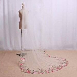 May include: A sheer, ivory wedding veil with a floral embroidered border. The embroidery features pink and red roses with green leaves. The veil is displayed on a mannequin, showcasing its length and delicate design, perfect for bridal wear.