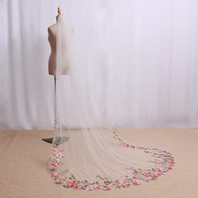 May include: A sheer, ivory wedding veil with a floral embroidered border. The embroidery features pink and red roses with green leaves. The veil is displayed on a mannequin, showcasing its length and delicate design, perfect for bridal wear.
