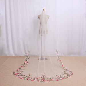 May include: A sheer, ivory-colored wedding veil with floral embroidery. The veil is displayed on a mannequin, showcasing its length and delicate design. The floral embroidery features pink and red flowers with green leaves.