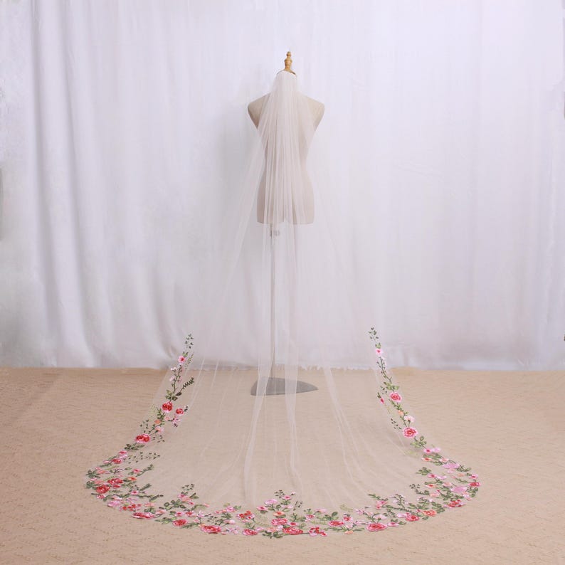 May include: A sheer, ivory-colored wedding veil with floral embroidery. The veil is displayed on a mannequin, showcasing its length and delicate design. The floral embroidery features pink and red flowers with green leaves.