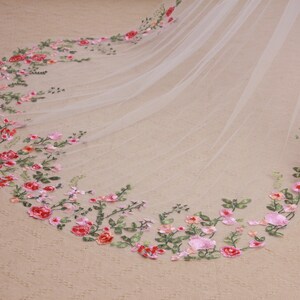 May include: A sheer, off-white wedding veil with an embroidered floral design. The flowers are in shades of red and pink, with green leaves. The veil is likely made of tulle or a similar lightweight fabric.