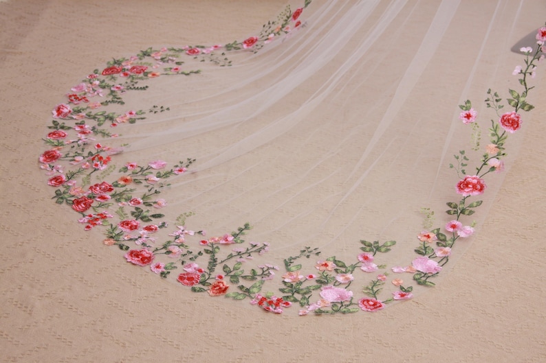 May include: A sheer, off-white wedding veil with an embroidered floral design. The flowers are in shades of red and pink, with green leaves. The veil is likely made of tulle or a similar lightweight fabric.