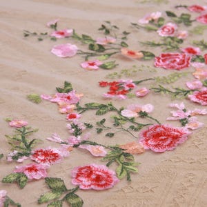 May include: Close-up of embroidered floral fabric. The design features red, pink, and peach-colored flowers with green leaves. The flowers are scattered across a beige, textured background, creating a delicate and romantic aesthetic.