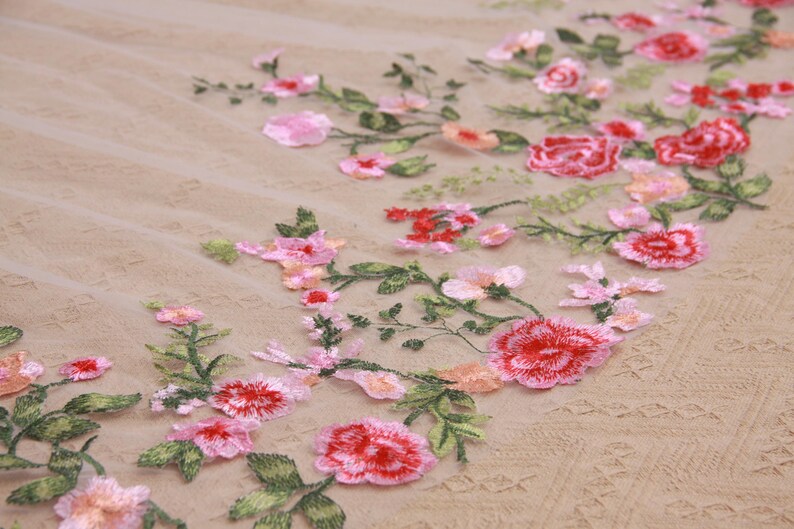 May include: Close-up of embroidered floral fabric. The design features red, pink, and peach-colored flowers with green leaves. The flowers are scattered across a beige, textured background, creating a delicate and romantic aesthetic.