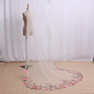 May include: A long, sheer white bridal veil with a floral embroidered border. The veil is displayed on a mannequin, showcasing the delicate fabric and the intricate design of pink and green flowers.