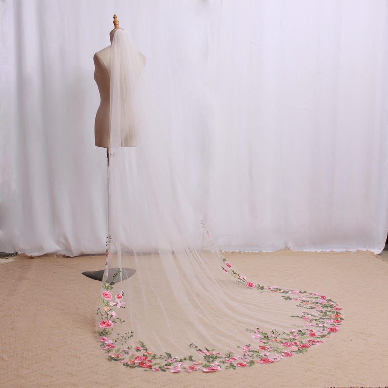 May include: A long, sheer white bridal veil with a floral embroidered border. The veil is displayed on a mannequin, showcasing the delicate fabric and the intricate design of pink and green flowers.