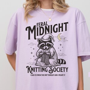 Puede incluir: Camiseta color orqu&iacute;dea con un gr&aacute;fico de un mapache tejiendo. El mapache lleva un gorro de dormir y una capa, con las palabras "Feral Midnight Knitting Society" arriba y abajo de la imagen. El texto "I said I'd finish this WIP tonight and I meant it" est&aacute; debajo.