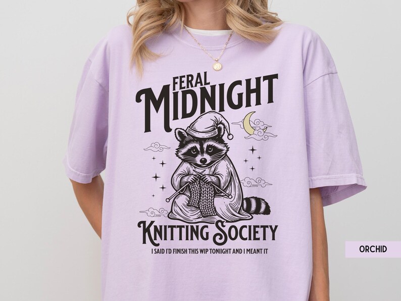 Puede incluir: Camiseta color orqu&iacute;dea con un gr&aacute;fico de un mapache tejiendo. El mapache lleva un gorro de dormir y una capa, con las palabras "Feral Midnight Knitting Society" arriba y abajo de la imagen. El texto "I said I'd finish this WIP tonight and I meant it" est&aacute; debajo.