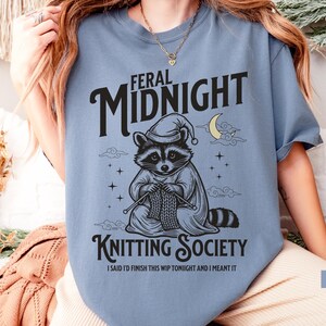 Puede incluir: Camiseta azul vaquero con un mapache con gorro de dormir y capa, tejiendo. La camiseta presenta el texto "FERAL MIDNIGHT KNITTING SOCIETY" con la frase "I SAID I'D FINISH THIS WIP TONIGHT AND I MEANT IT".