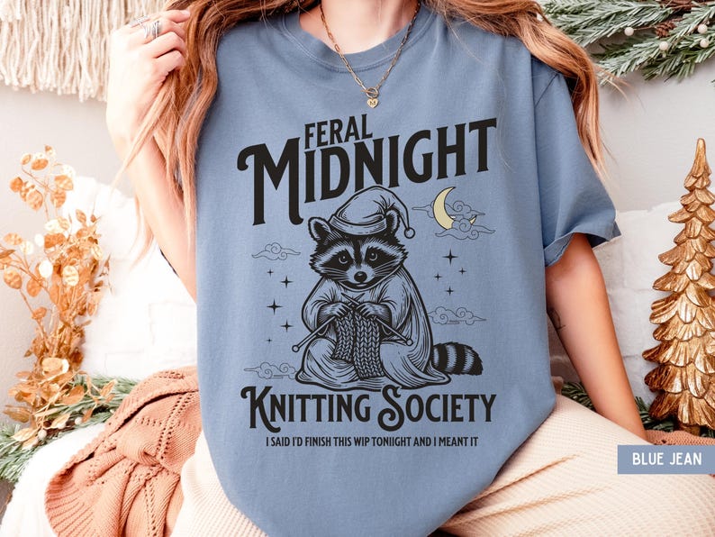 Puede incluir: Camiseta azul vaquero con un mapache con gorro de dormir y capa, tejiendo. La camiseta presenta el texto "FERAL MIDNIGHT KNITTING SOCIETY" con la frase "I SAID I'D FINISH THIS WIP TONIGHT AND I MEANT IT".